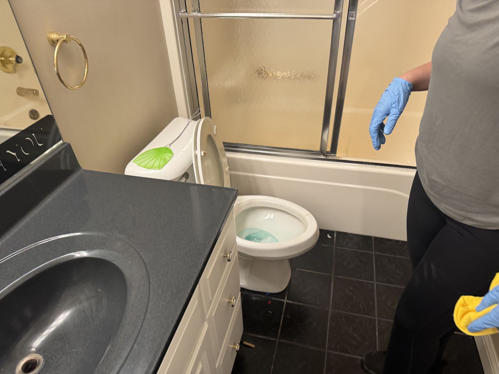 A person in blue gloves and grey clothing stands in a bathroom, holding a yellow cleaning cloth near a sink and toilet.