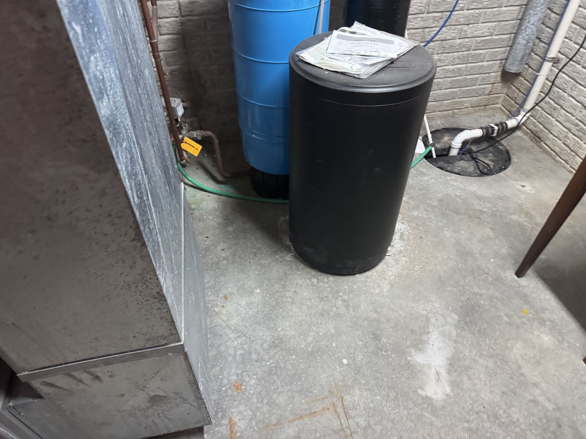 A blue water treatment tank and a black salt brine tank sit on a concrete floor in a utility or basement area.