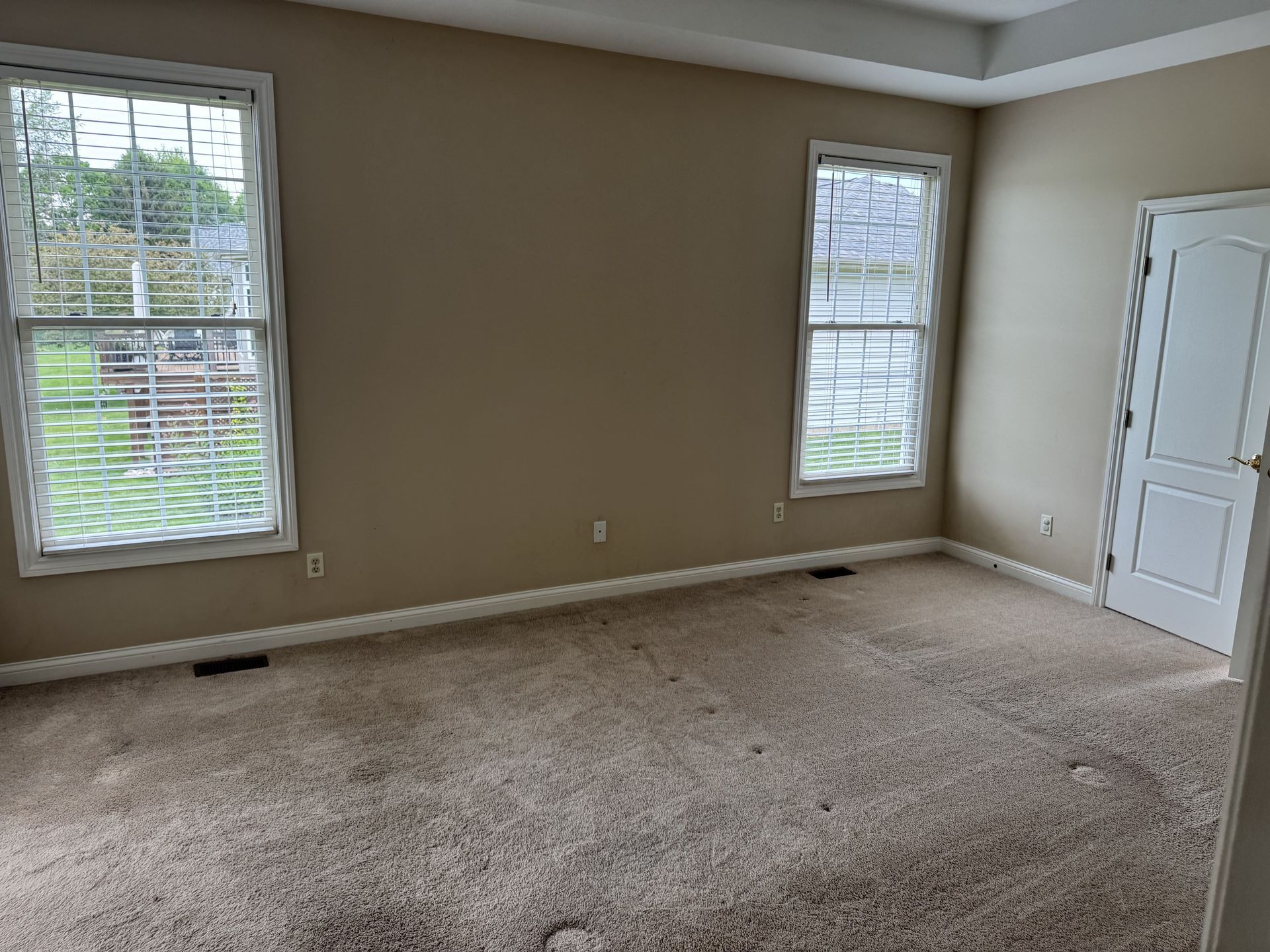 An empty room with tan walls, light carpet, two windows with blinds, and a white door on the right.