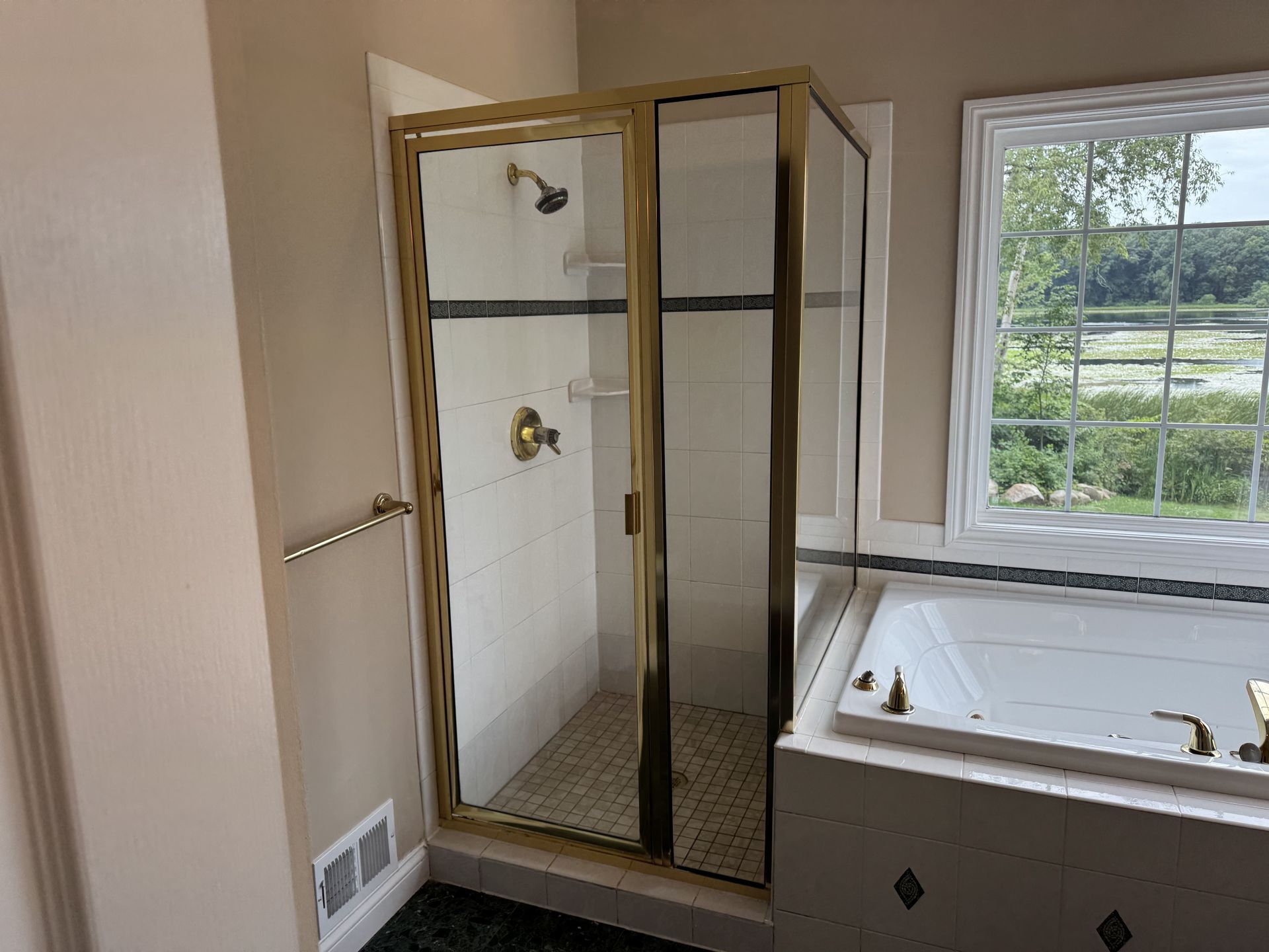 A bathroom featuring a gold-framed glass shower stall next to a white tiled bathtub, with a window overlooking greenery.