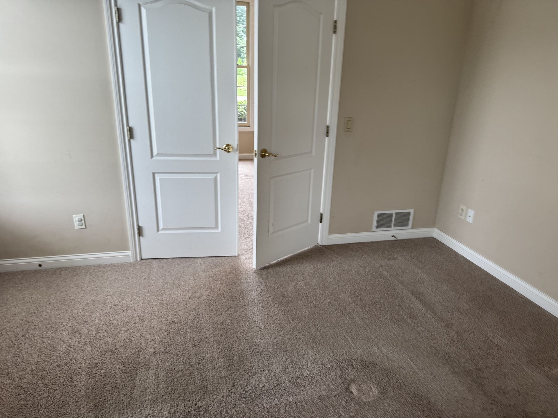 A carpeted room with beige walls, featuring double white doors slightly ajar, revealing a view into another room.