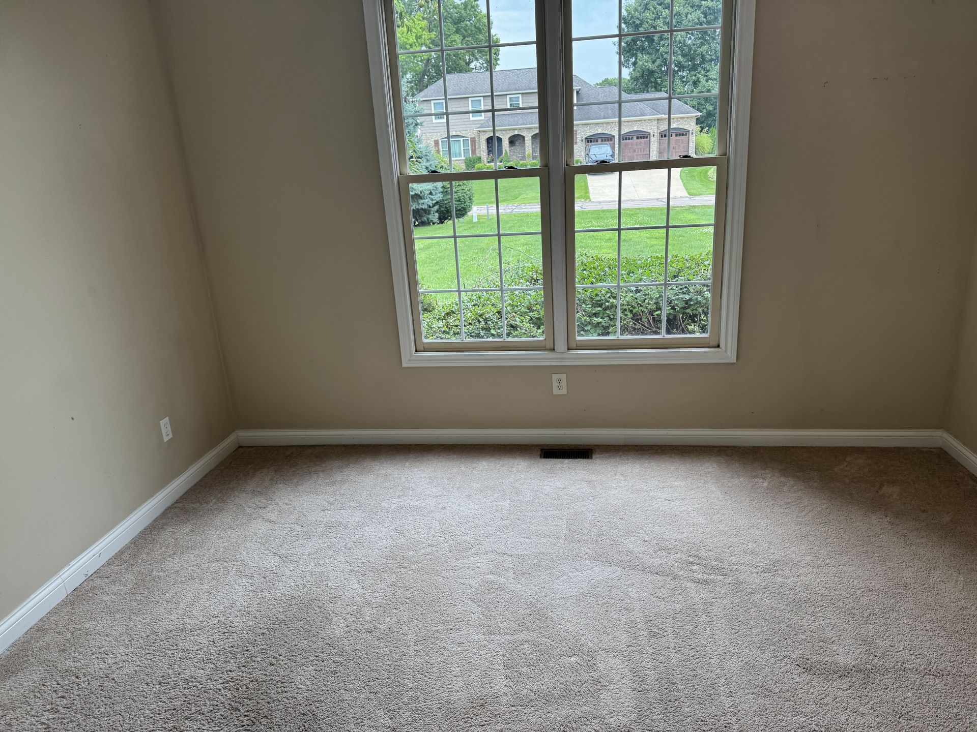 An empty room with beige walls, tan patterned carpet, white baseboards, and a large window looking out onto a lawn.