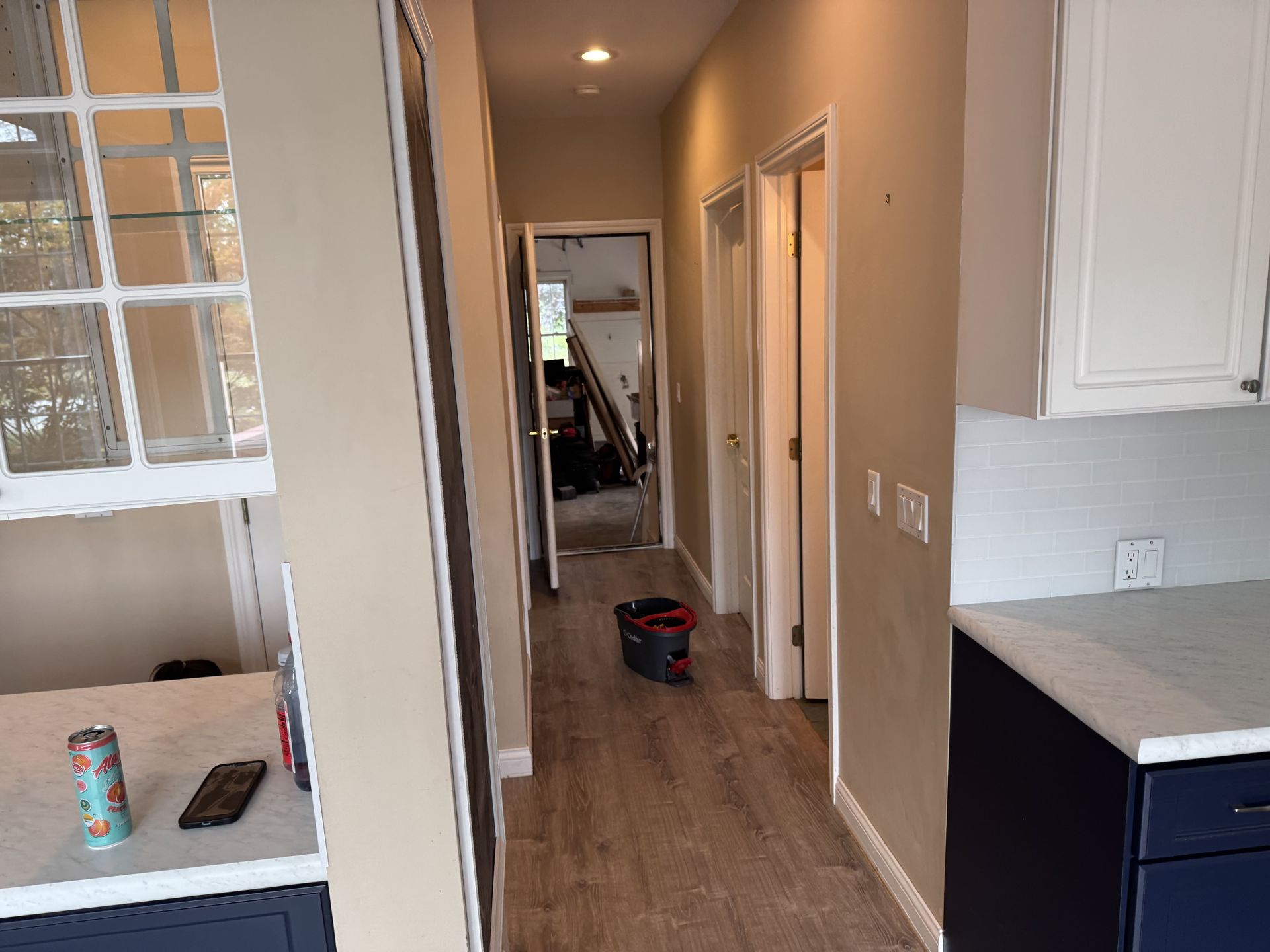A narrow hallway with light wood flooring leads to an open doorway, flanked by kitchen counters on both sides.