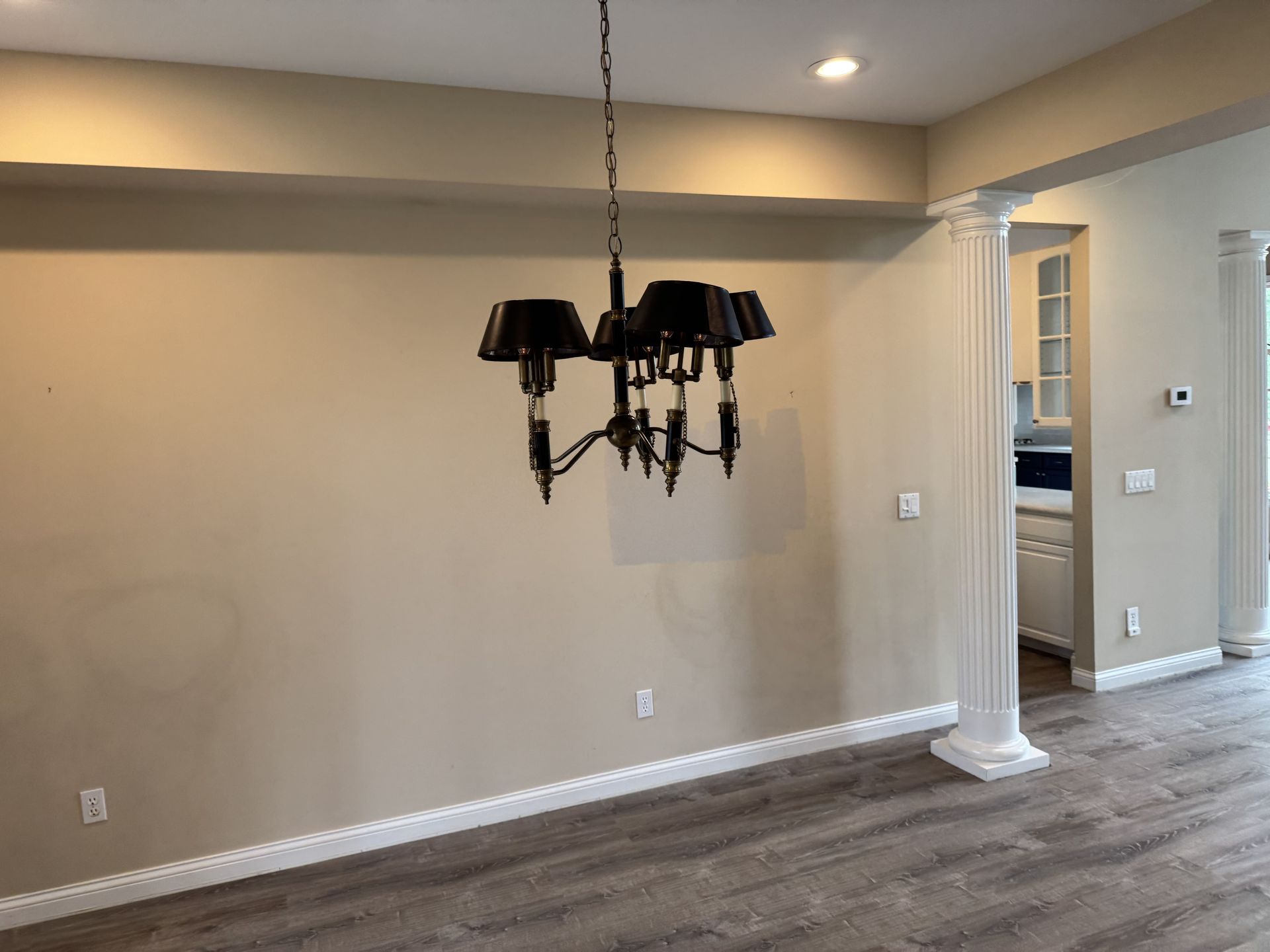 A black chandelier hangs in a neutral-toned room with wood-look flooring and a white column leading to a kitchen.