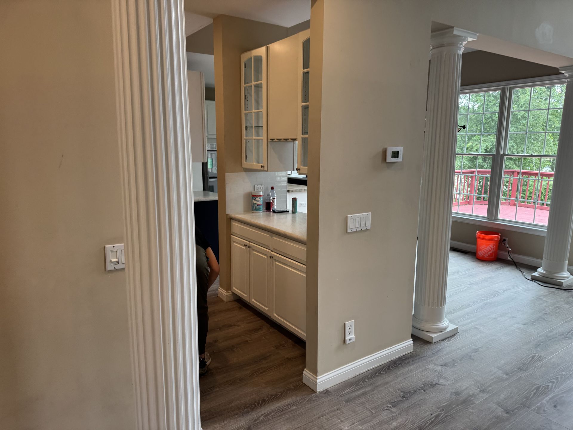 A beige room with white columns, light wood flooring, a kitchen area with white cabinets, and windows looking outside.