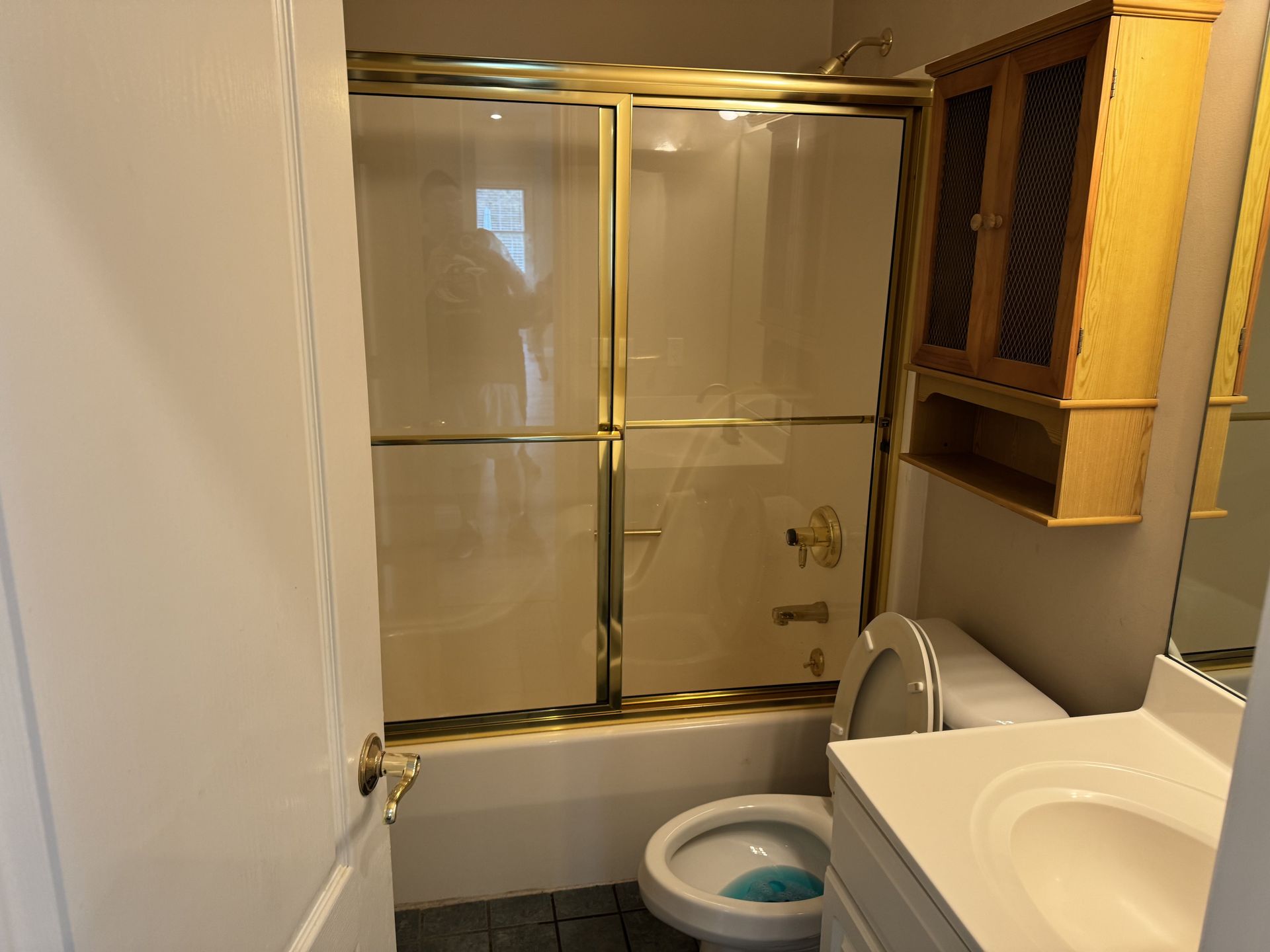 A small bathroom with a bathtub, sliding glass shower door, toilet, vanity sink, and a wooden wall cabinet.