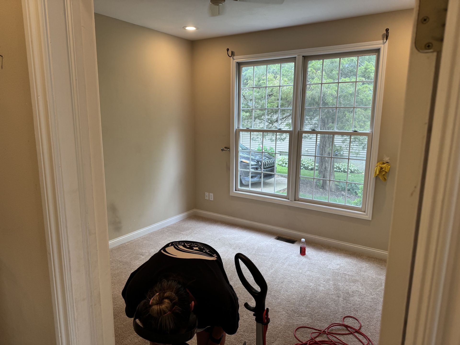 A person seen from behind vacuums the beige carpet of an empty, light-filled bedroom with a large window.