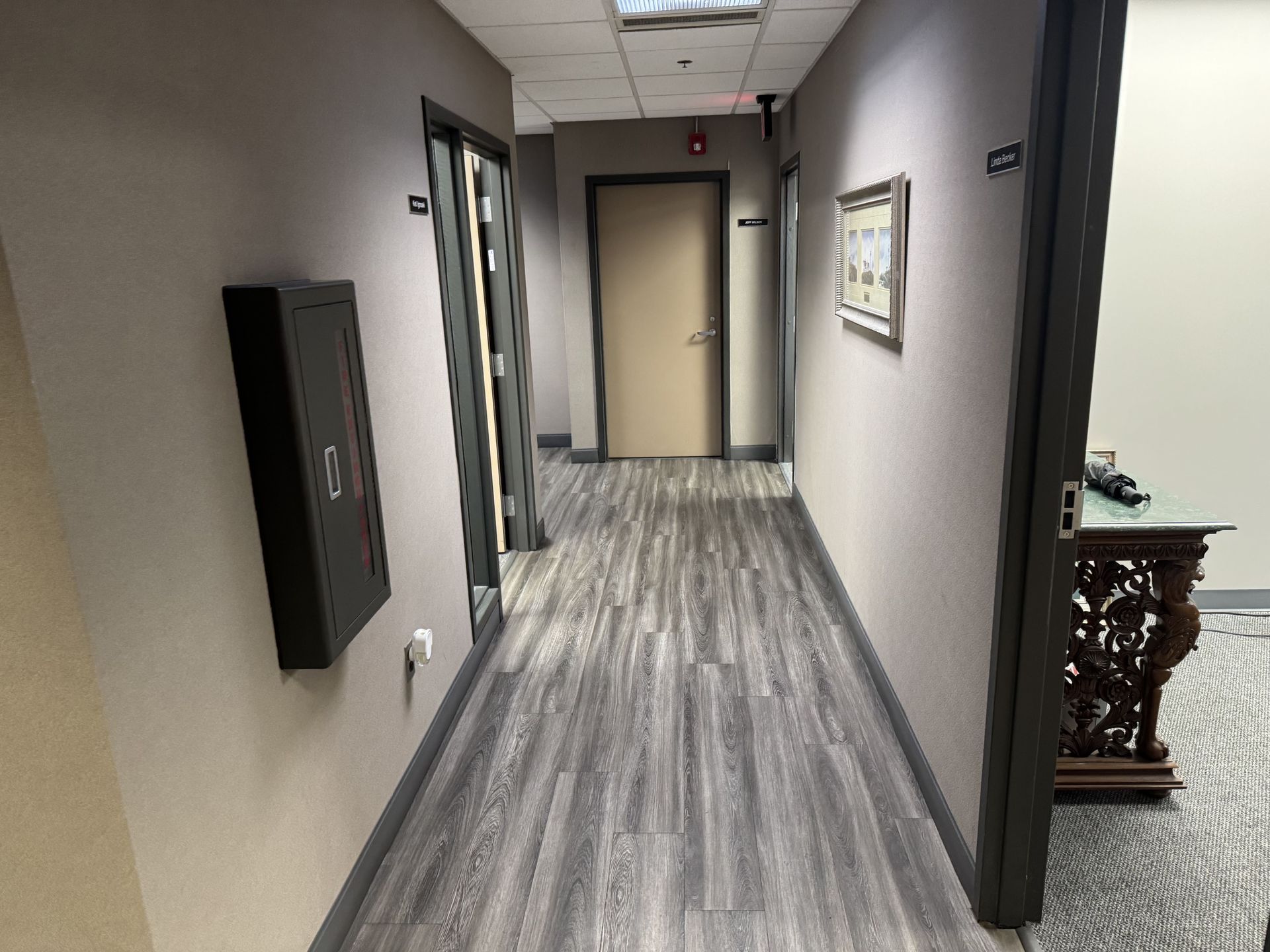 A view down a neutral-toned office hallway with grey wood-patterned flooring, several doors, and a wall-mounted cabinet.