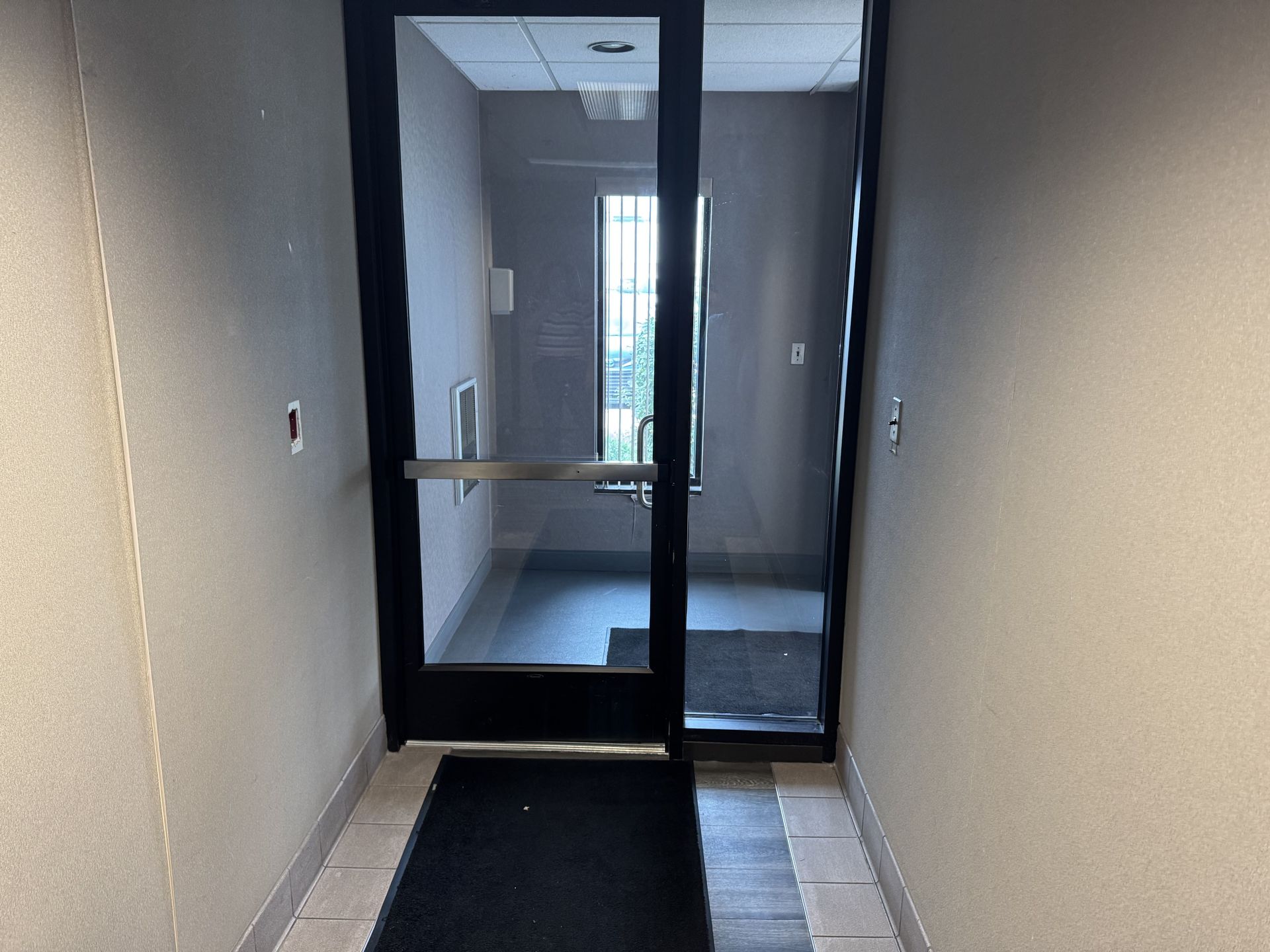 A narrow hallway leads to a glass door with a black frame and a black floor mat, showing a windowed room beyond.