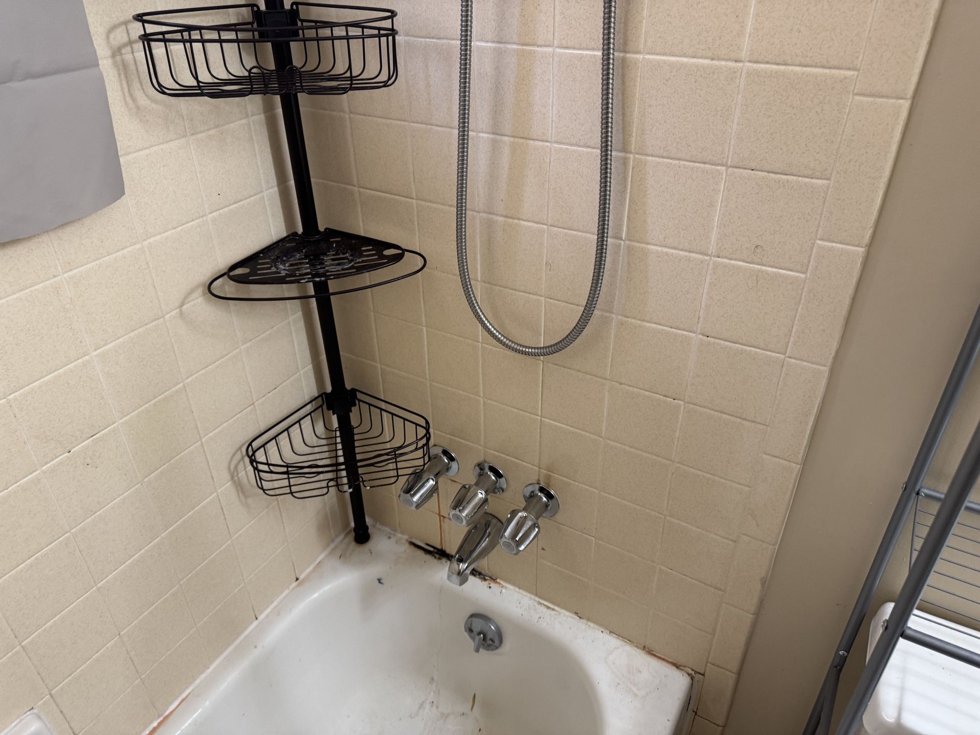 A white bathtub with a three-tier black corner shower caddy and a three-handle chrome faucet on a tiled wall.