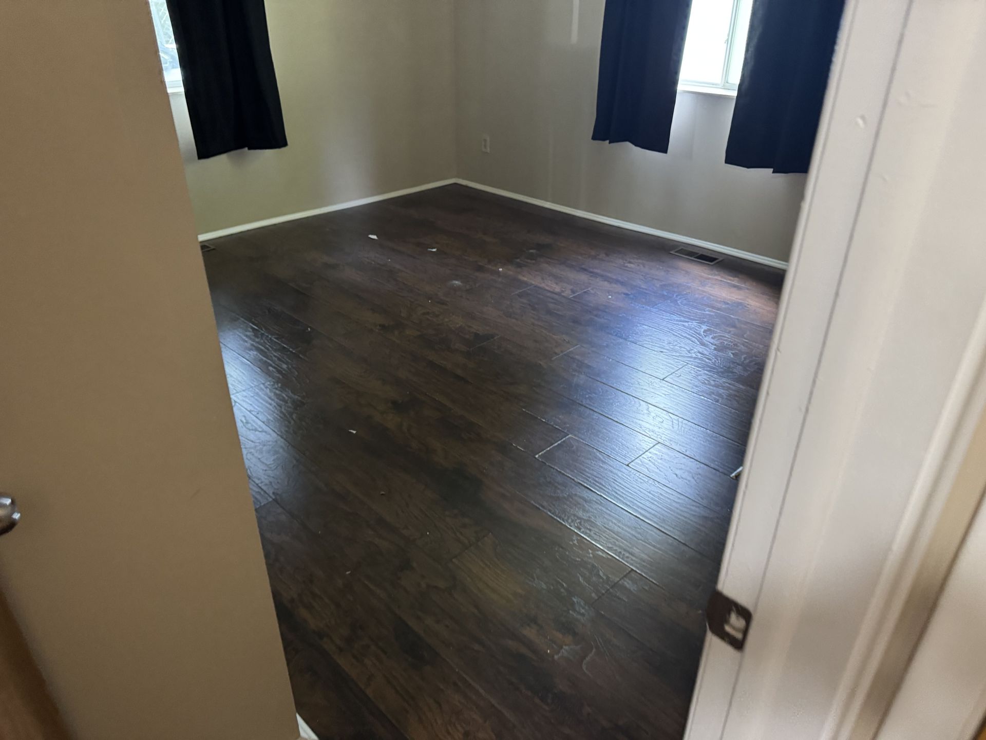 An empty room featuring dark wood-look laminate flooring, light gray walls, and black curtains over two windows.