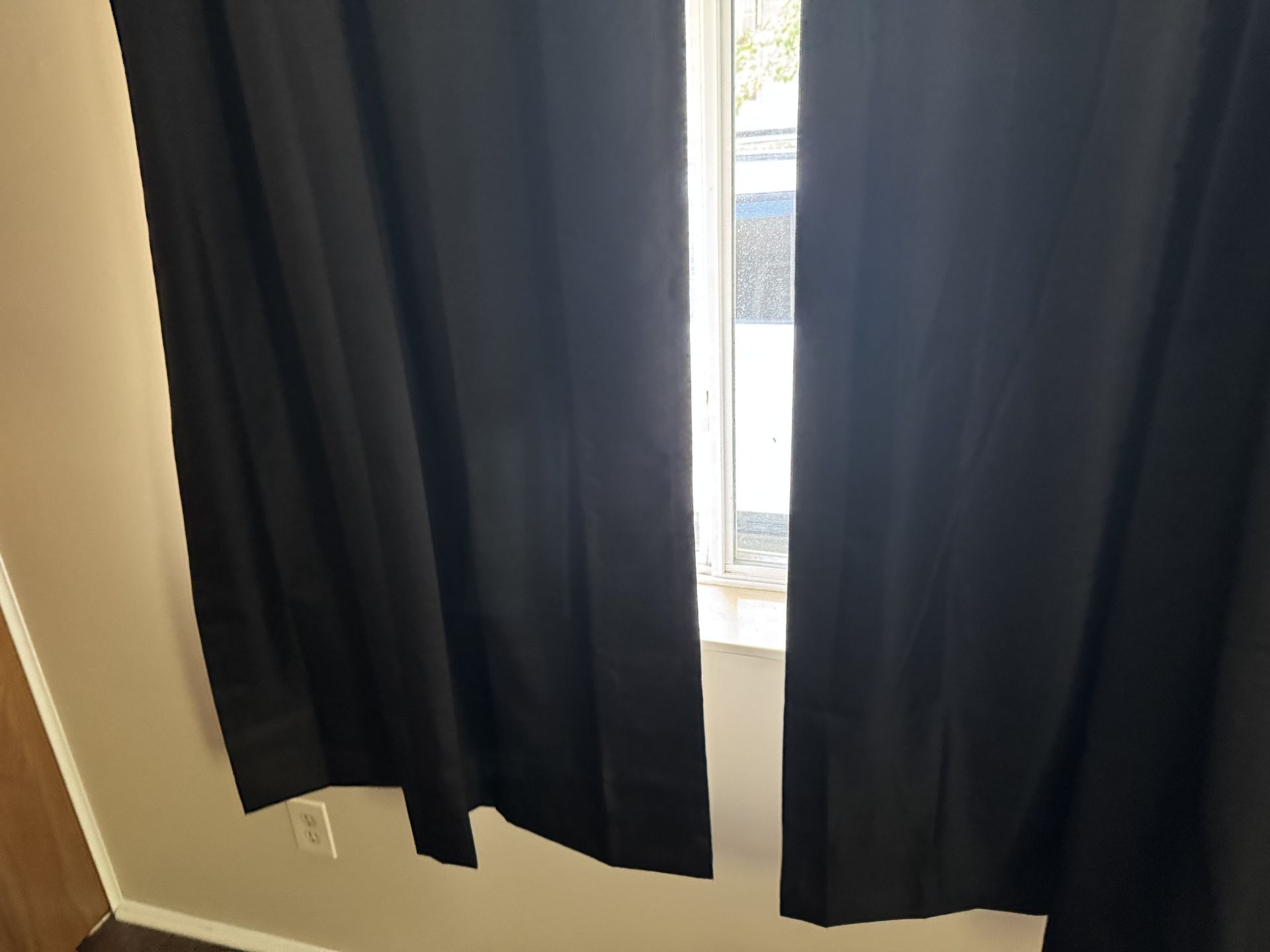 Black curtains hanging on a rod over a window, with a small gap in the center revealing a view outside.