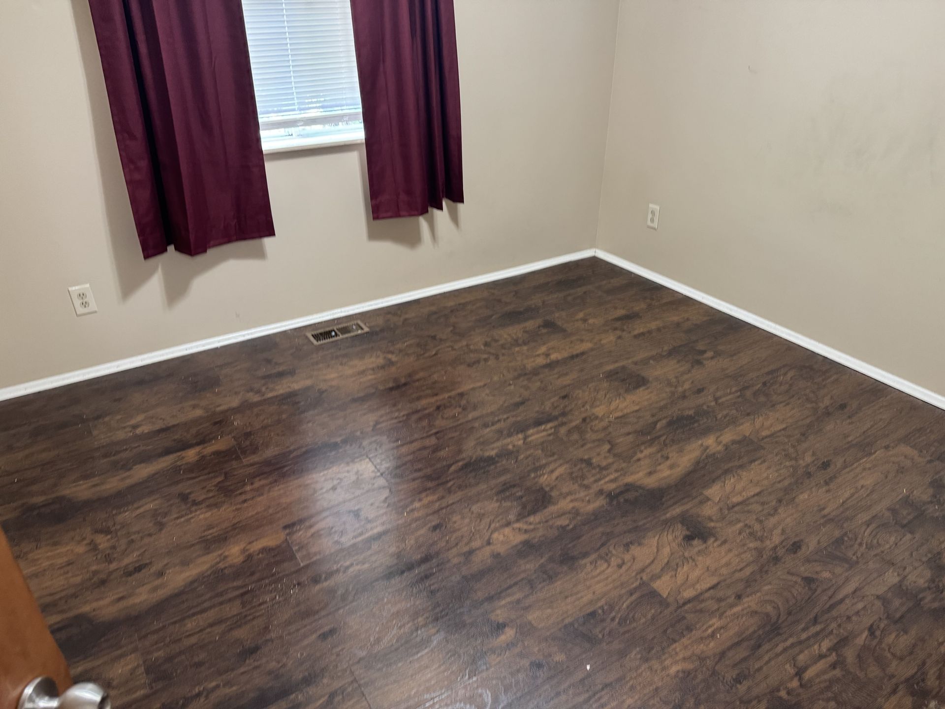An empty room with dark wood-look laminate flooring, beige walls, and maroon curtains hanging over a small window.