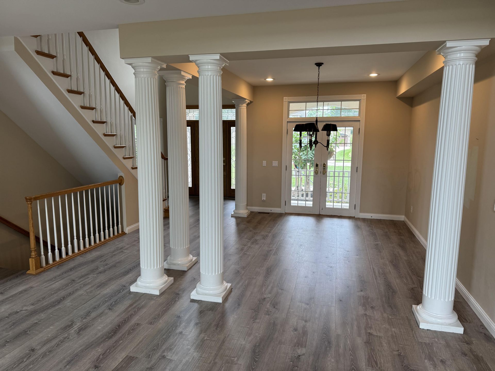 A light-filled entryway with grey wood-look floors, a staircase with a wooden railing, and several white fluted columns.
