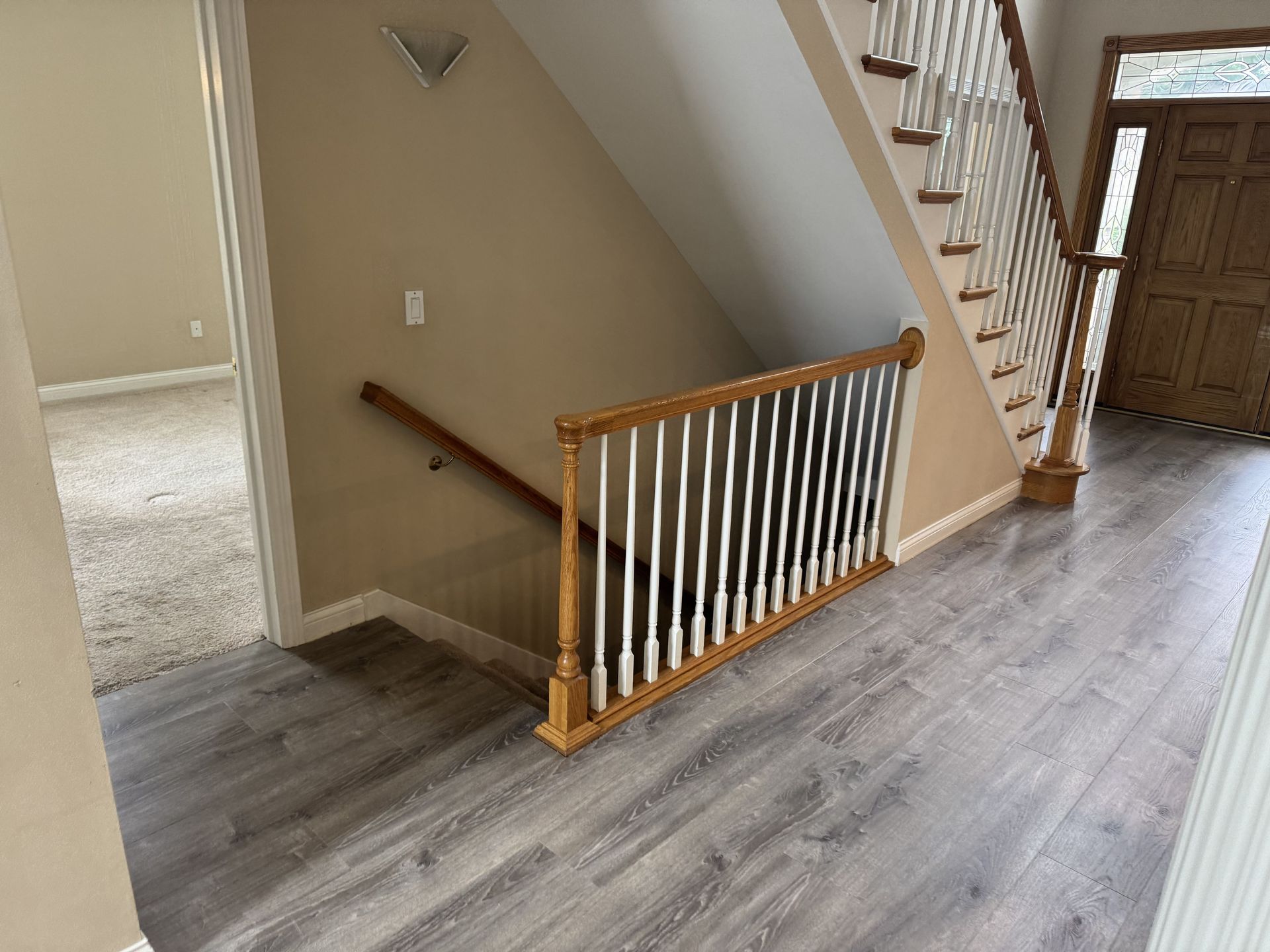 A view from a hallway showing light wood-look floors, a wooden staircase with white balusters, and an entryway door.