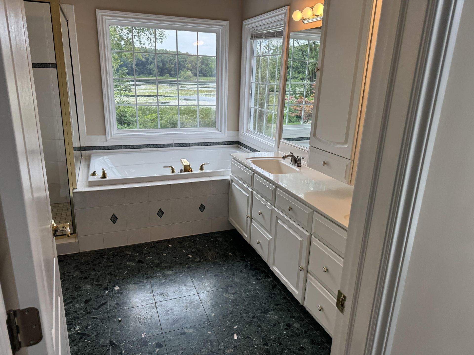 A bathroom featuring a white soaking tub by a large window, a double-sink vanity, and dark green speckled flooring.