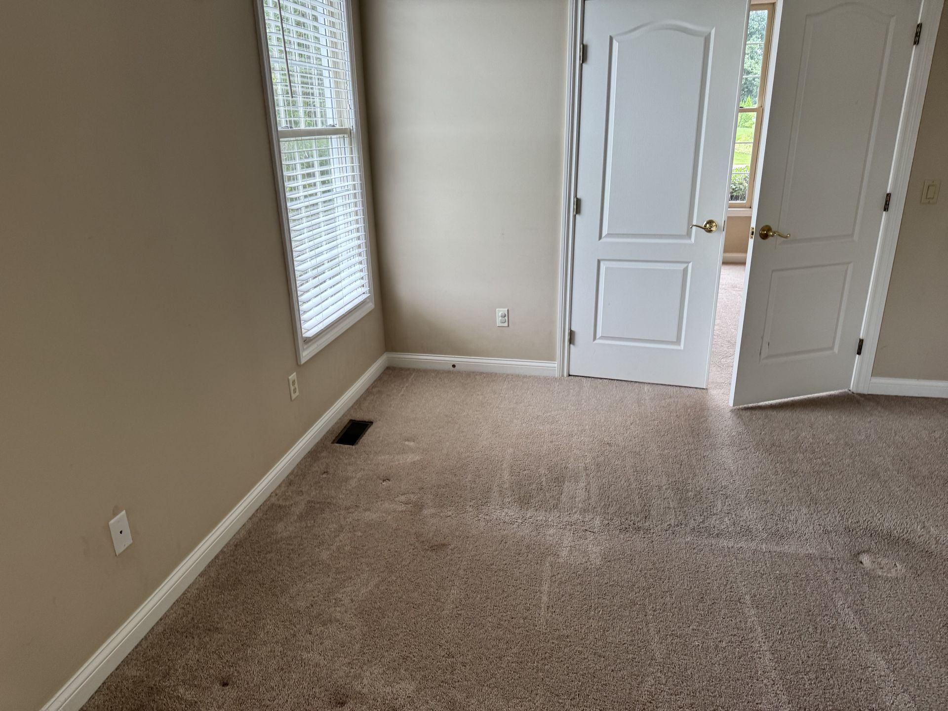 An empty room with beige walls, brown carpet, a window with blinds, and white double doors opening to another room.