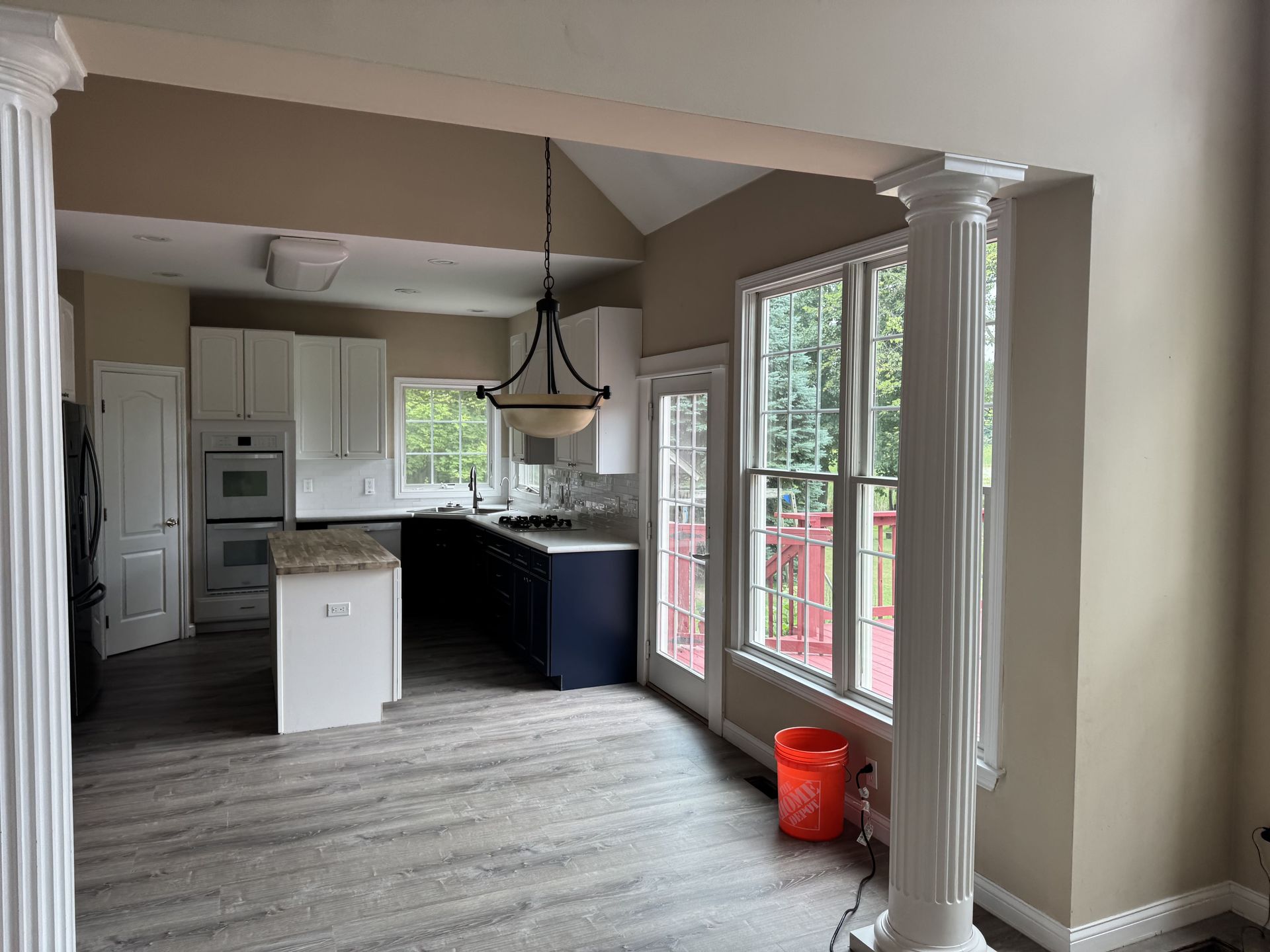 A kitchen with a white island, dark cabinets, and light wood flooring, viewed through two white decorative pillars.
