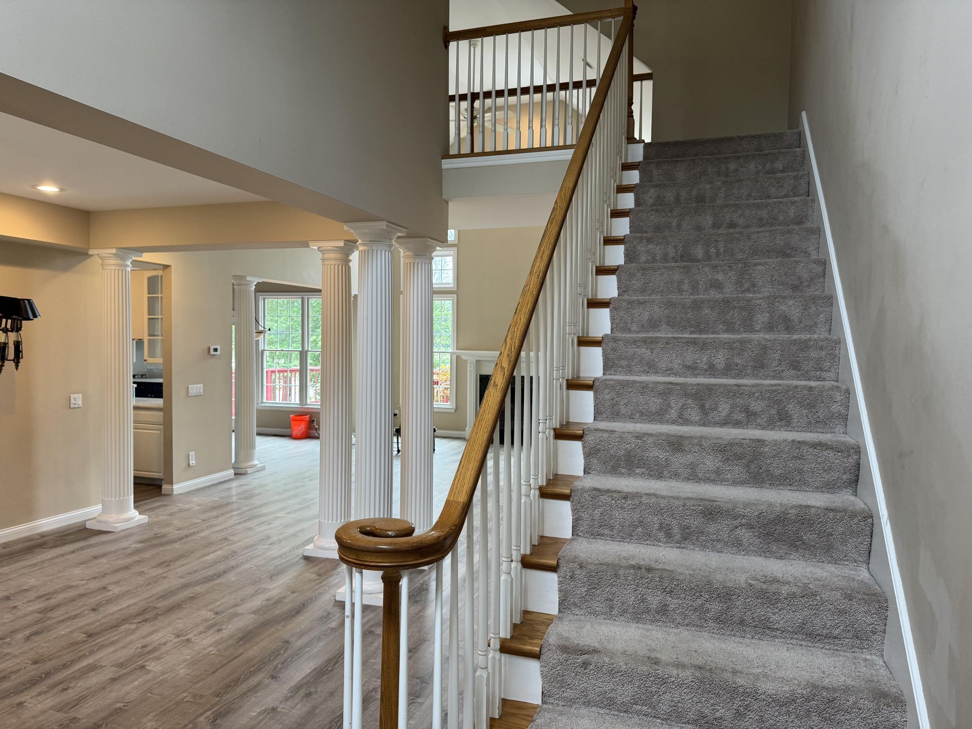 Carpeted staircase with white railings leading up, next to an open floor plan room with light wood floors and white columns.