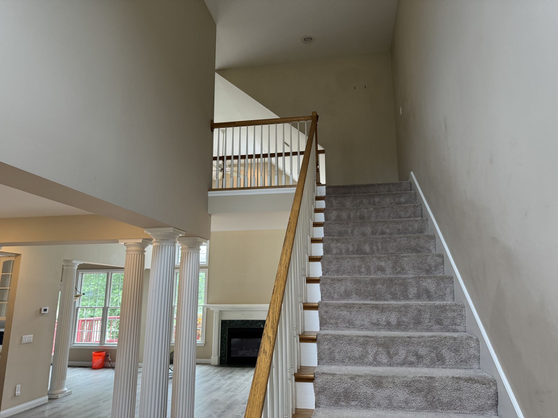 A carpeted staircase with a wooden railing leads up to an open landing in a house with high ceilings and decorative columns.