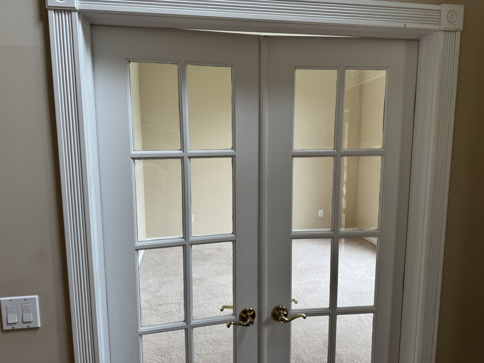 White French doors with eight glass panes each, set in a textured white frame against a tan wall with a light switch.