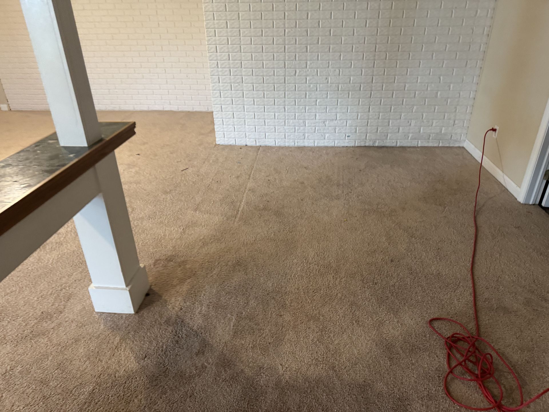 A room with tan wall-to-wall carpeting, a white brick wall, a white support pillar, and a red electrical extension cord.