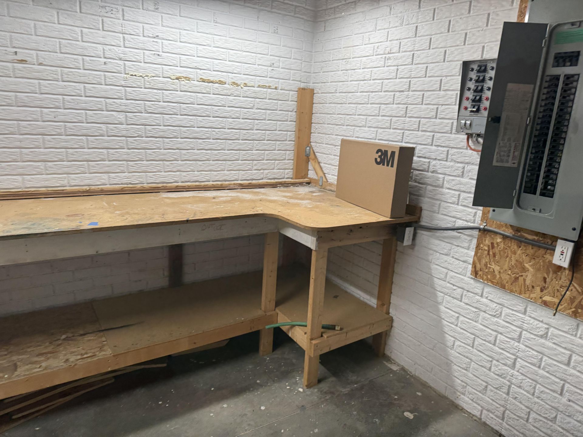A wooden workbench against a white brick wall next to an open electrical panel.
