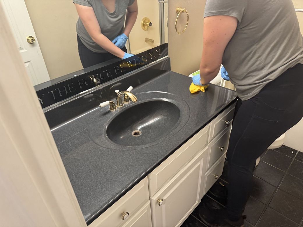 Person cleaning a bathroom sink and countertop with a yellow cloth in front of a mirror.