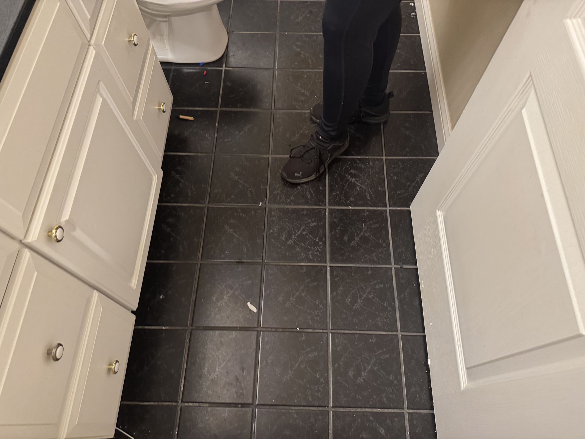 A person stands on a black tiled floor in a bathroom between white cabinets and an open white door.