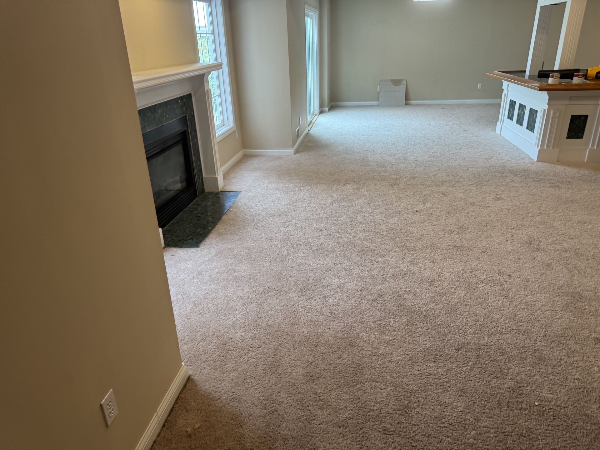 A spacious room featuring beige carpet, a fireplace with a dark hearth, and a white built-in bar counter in the distance.