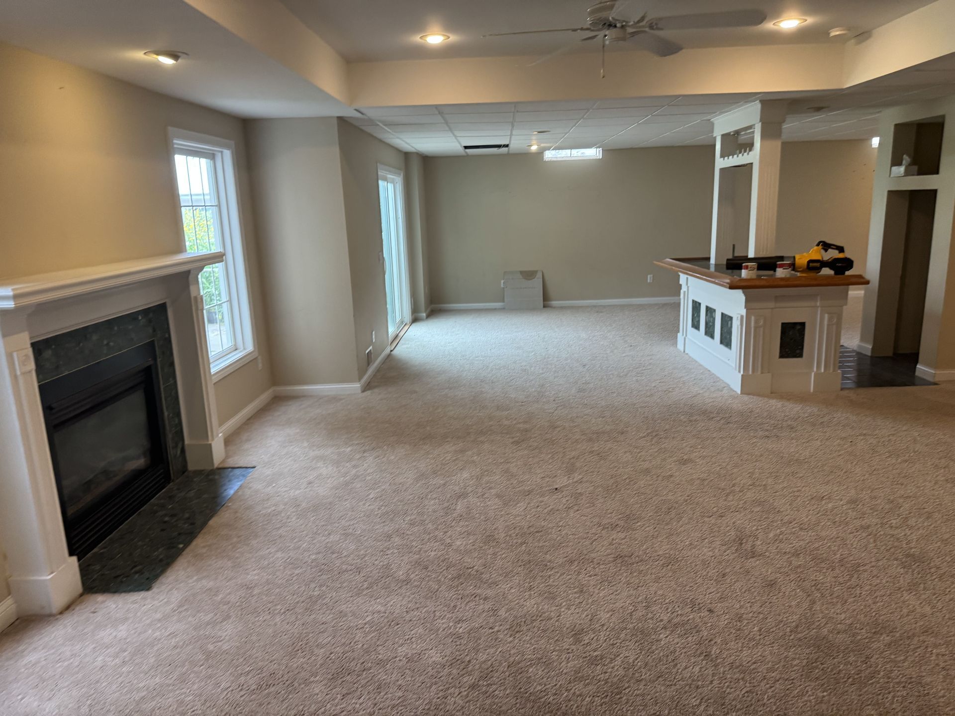 A wide-angle view of a beige basement featuring a fireplace, tan carpet, a wet bar, and recessed lighting.