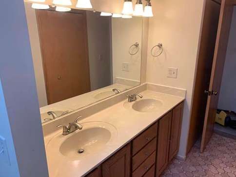 A double-sink bathroom vanity with light-colored countertops, wood cabinets, a large mirror, and overhead light fixtures.