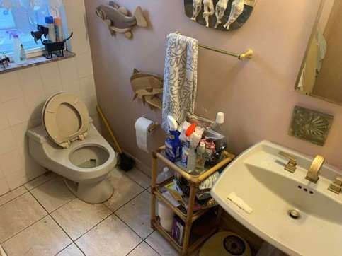 Small bathroom with light-colored tile floor, a wall-mounted toilet, sink, and a bamboo storage rack holding toiletries.