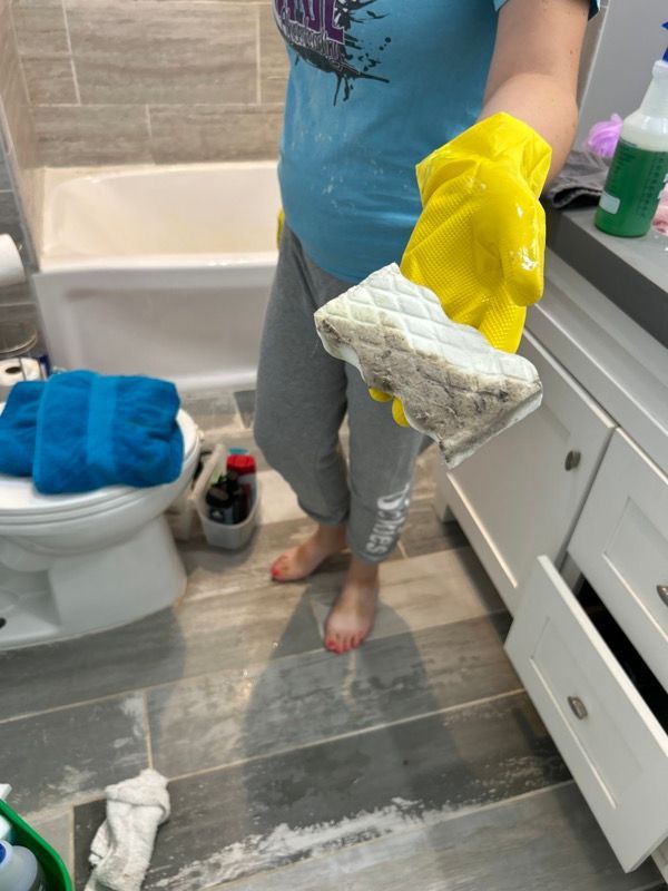 A person wearing a yellow rubber glove holds up a dirty, quilted cleaning sponge inside a tiled bathroom.