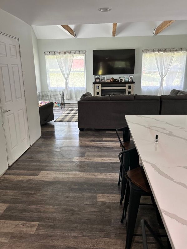 An open-concept room featuring a gray sectional sofa, a fireplace with a TV, exposed wood beams, and a marble kitchen island.