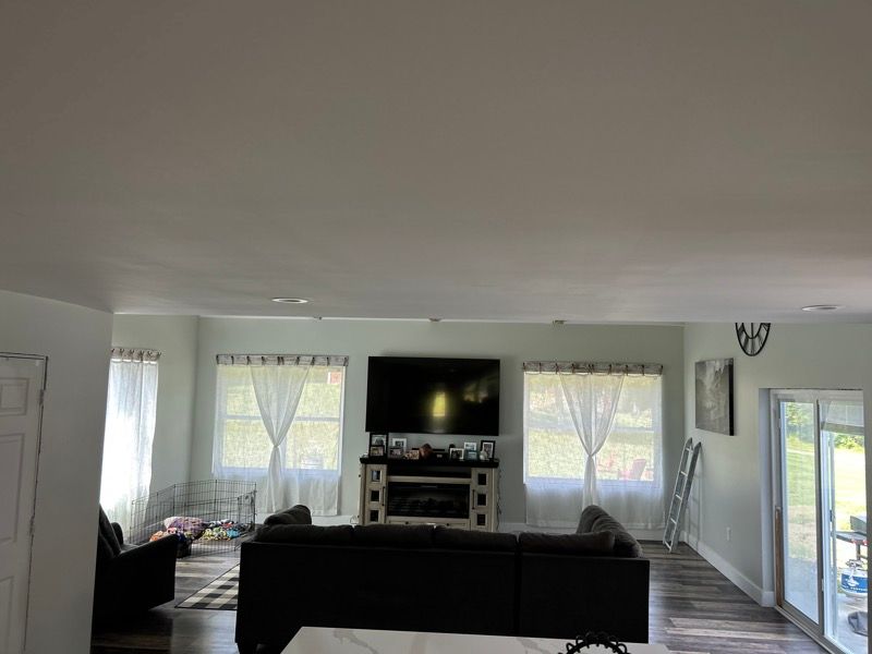 A modern, bright living room with a large gray sectional sofa, a fireplace with a TV above it, and wide windows with drapes.