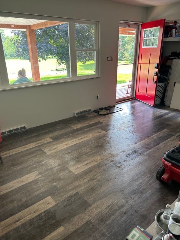 A room with wood-look plank flooring, a large picture window, and a bright red door leading to an outdoor porch.