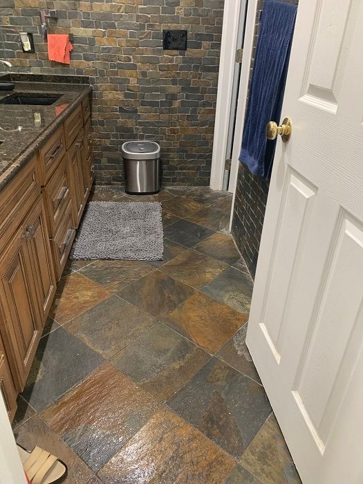 A bathroom with wood vanity cabinets, a stone tiled floor, and a stone-look wall with a trash can and hanging towel.