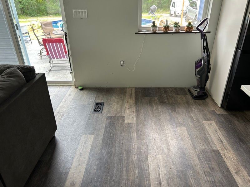 A light-filled room featuring wood-look vinyl plank flooring, a sliding glass door, a window sill with plants, and a vacuum.