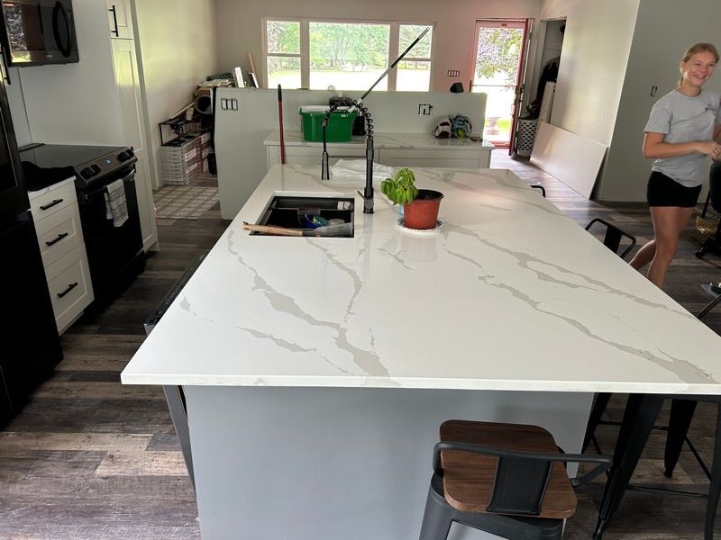 A person stands in a kitchen featuring a large white quartz island with gray veining, a black sink, and gray cabinetry.