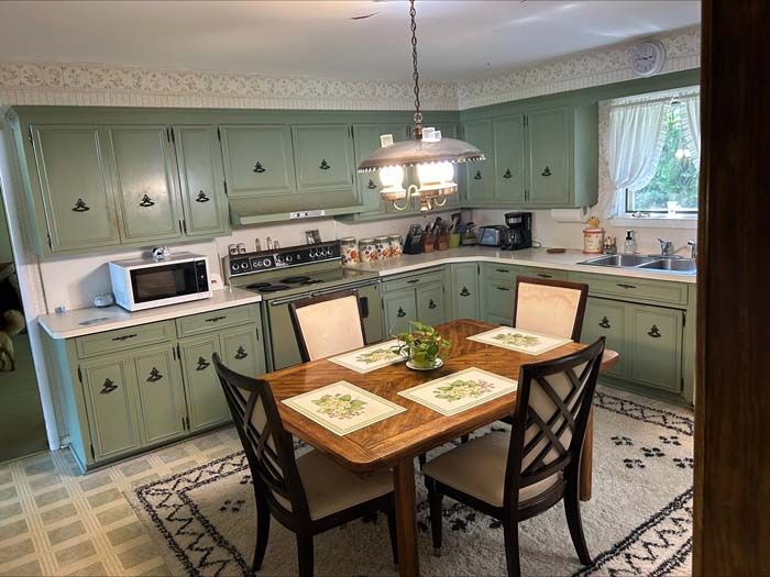 A kitchen featuring sage green cabinets, a wooden table with four chairs on a patterned rug, and a central light fixture.
