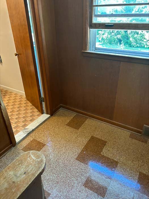 A room with light-colored, speckled vinyl tile flooring, wood-paneled walls, a single window, and an open wooden door.