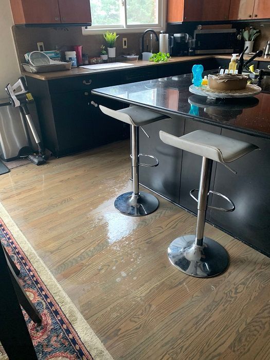 A kitchen with two grey bar stools and a spill of liquid on the wooden floor in front of a dark island.