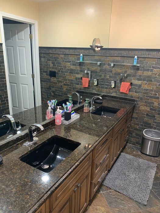 A double-sink bathroom vanity with dark brown countertops, black sinks, wood cabinets, and a slate-tiled wall.