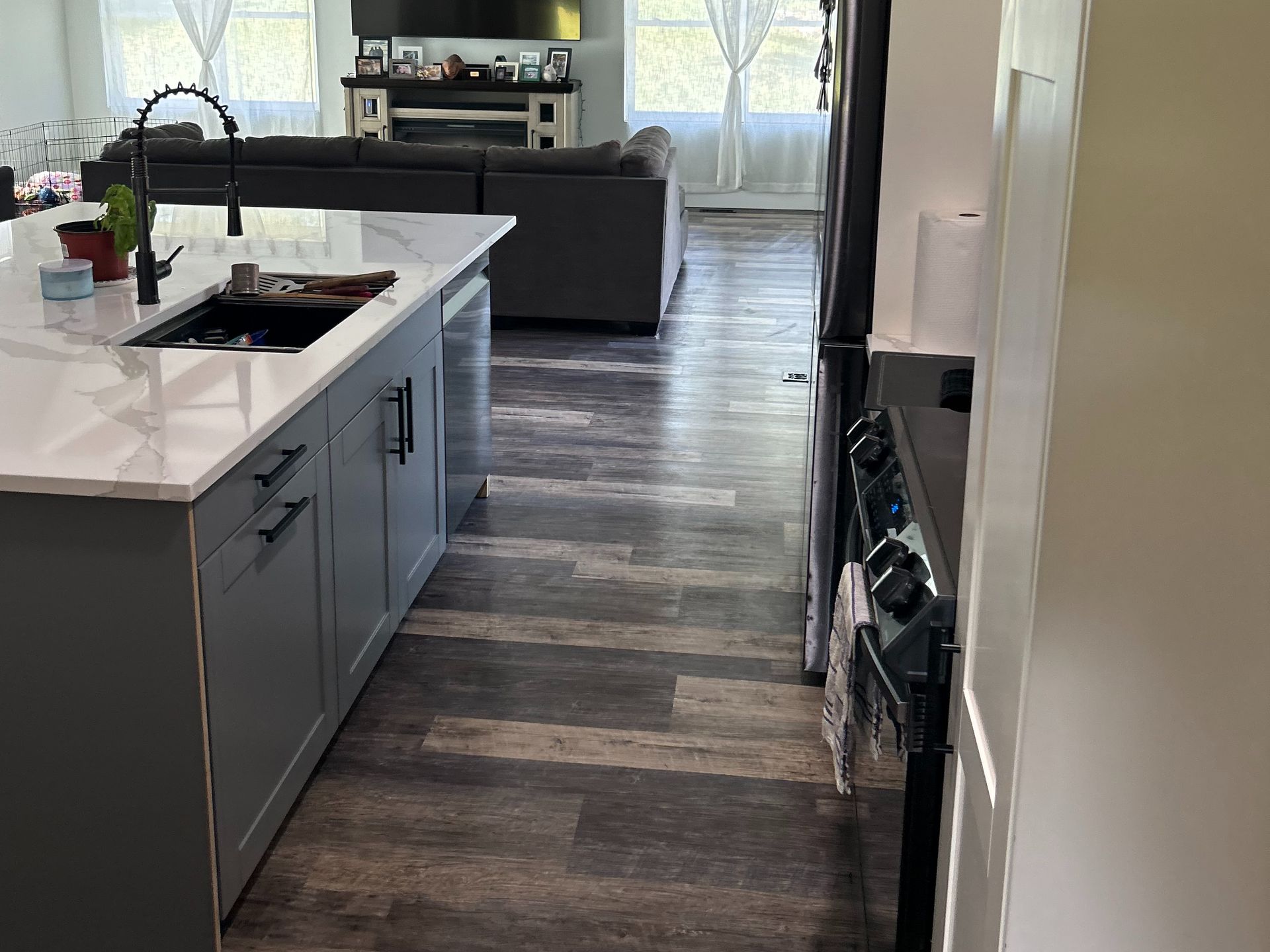 A modern kitchen with a gray island and sink in the foreground, leading to an open-plan living room with wood-style flooring.