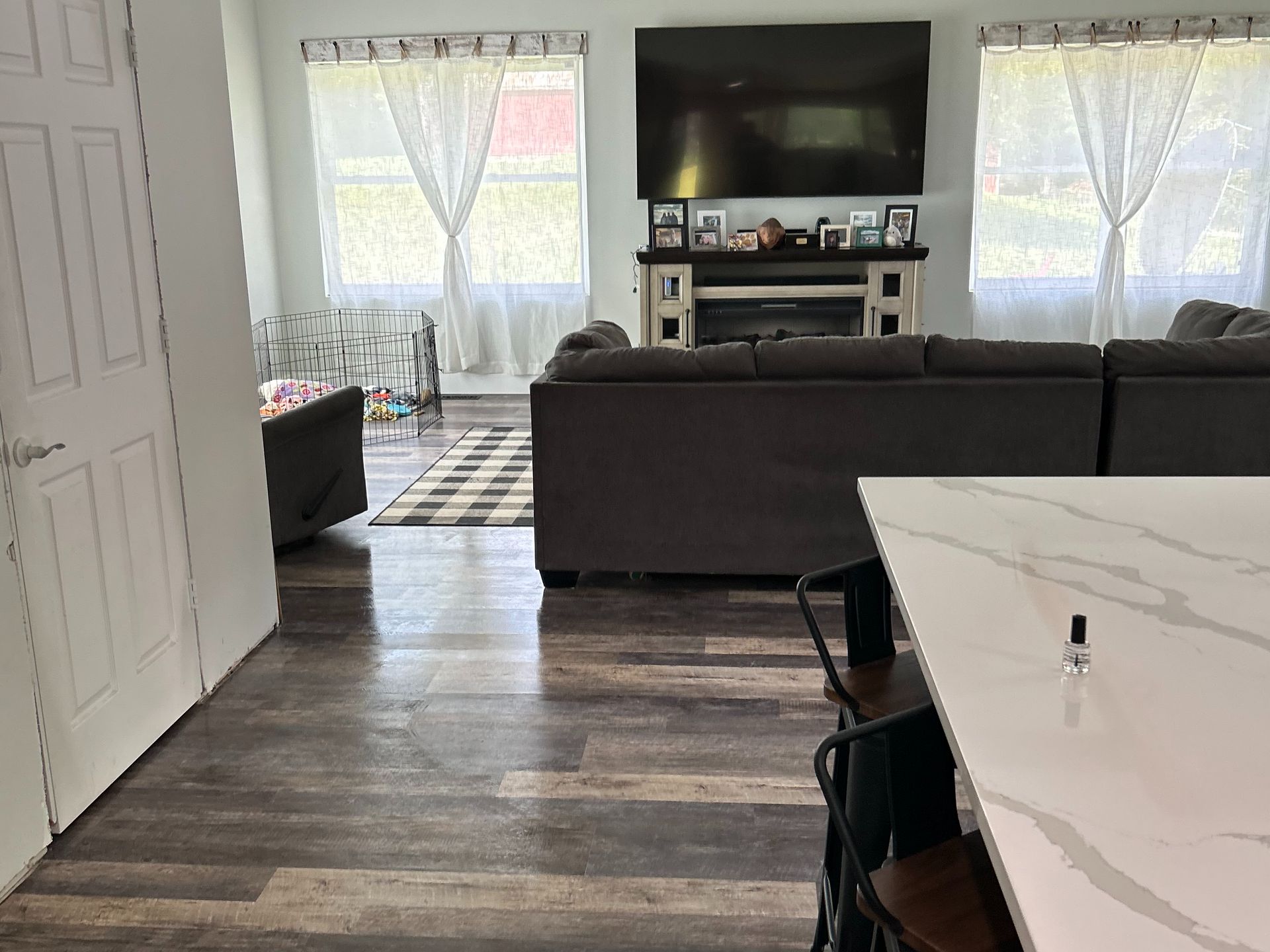 A view from a kitchen island overlooking a living room with a large sofa, fireplace, TV, and light-filtering curtains.