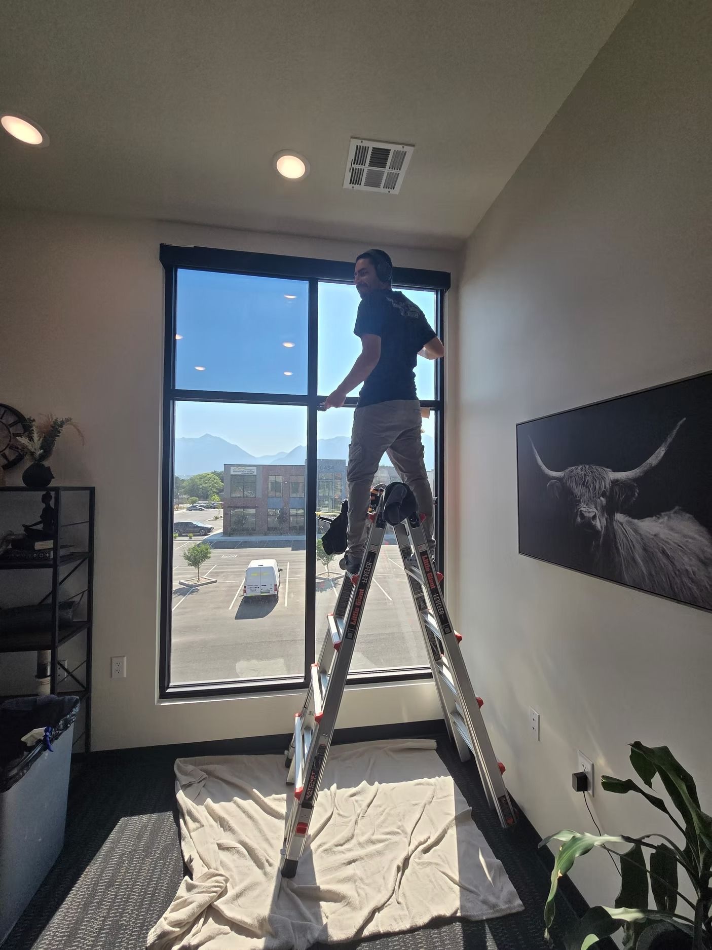 Man on ladder installing window film in a room with tall windows, with a mountain view.