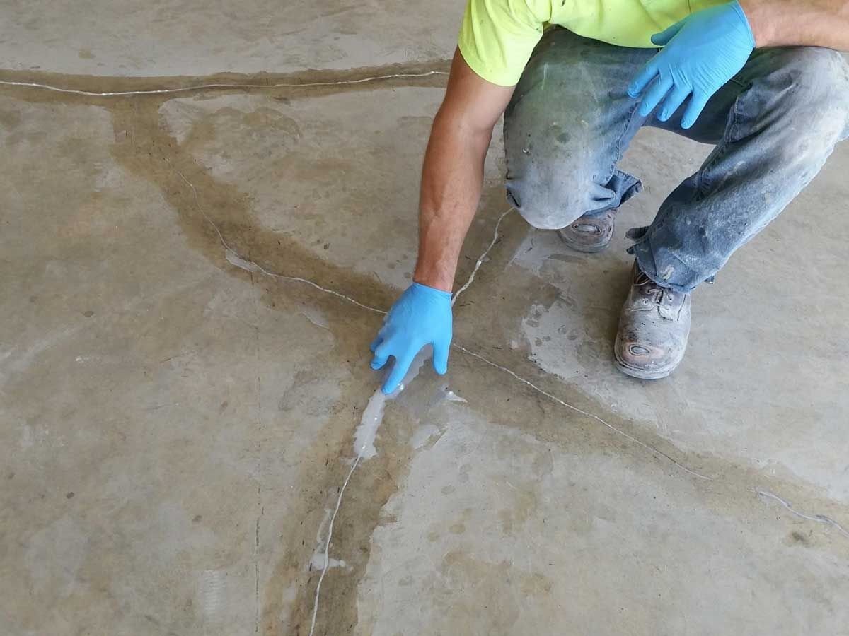 Person filling cracks in a concrete floor with a trowel, wearing gloves.