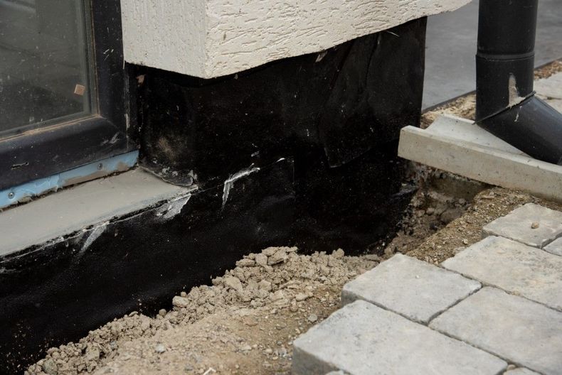 Damaged black wall base near a window and paving stones, with exposed dirt along the edge.