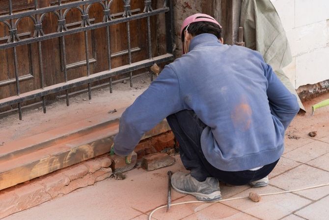 Person crouching by a building wall, repairing a damaged brick step with tools.